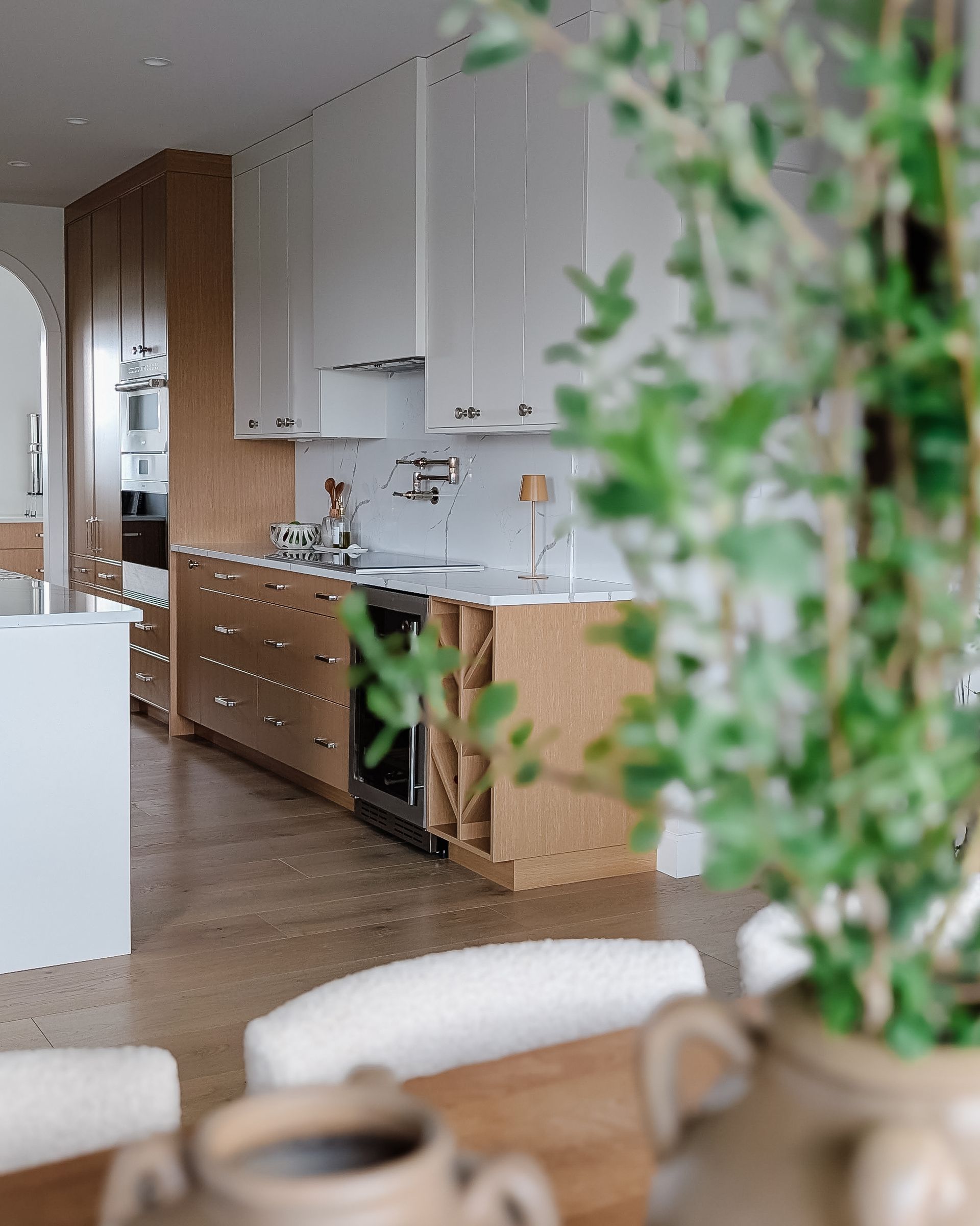 Modern kitchen with light wood and white cabinets, marble countertops, and a plant in foreground.