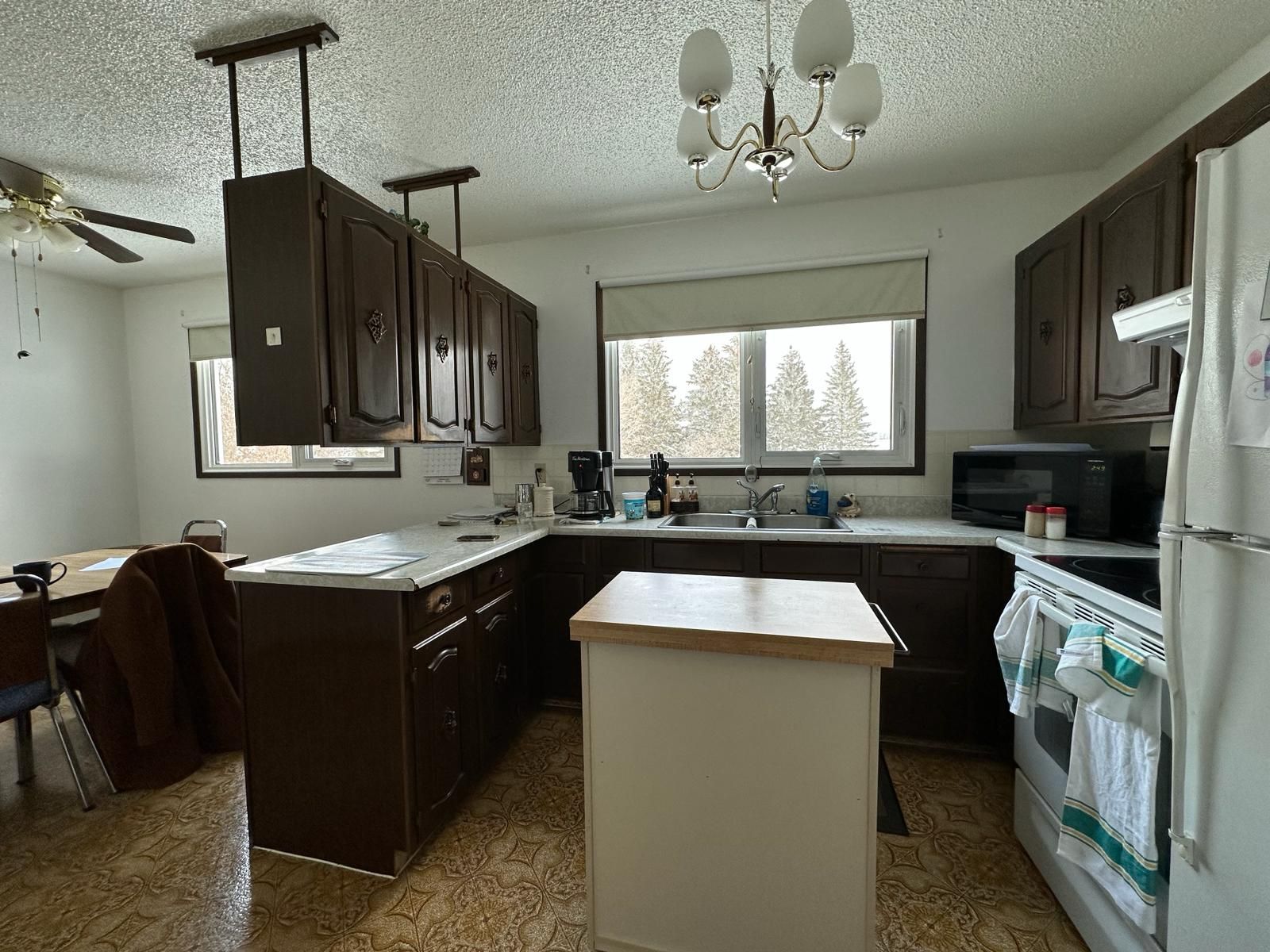 Kitchen with dark brown cabinets, white countertops, and small island. Window over sink, white appliances.