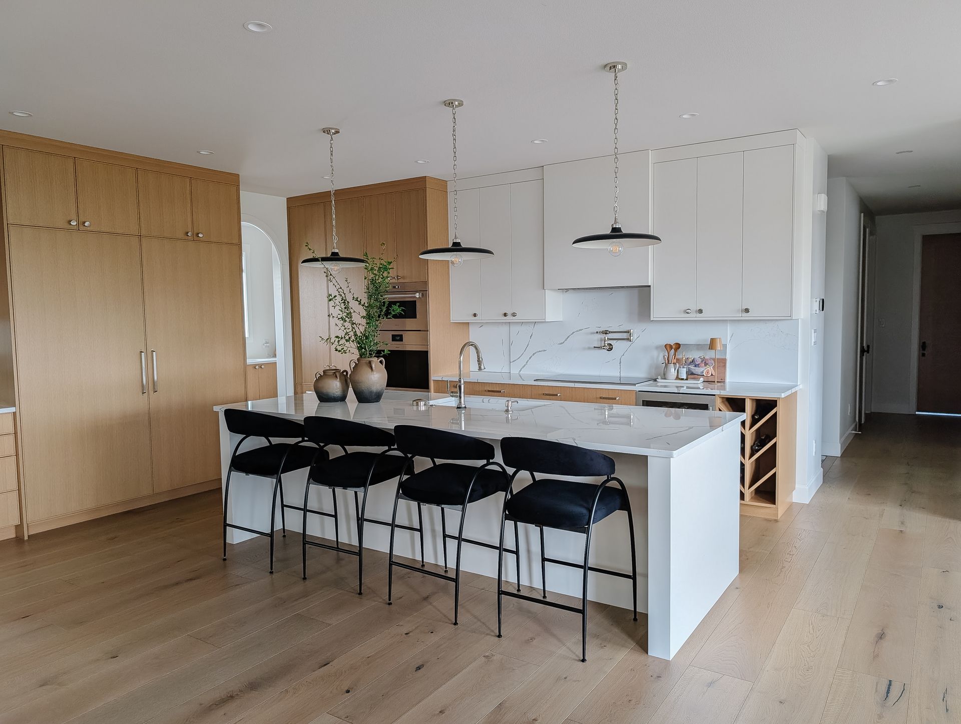 Modern kitchen with white and wood cabinets, island with black stools, pendant lights, and hardwood floors.