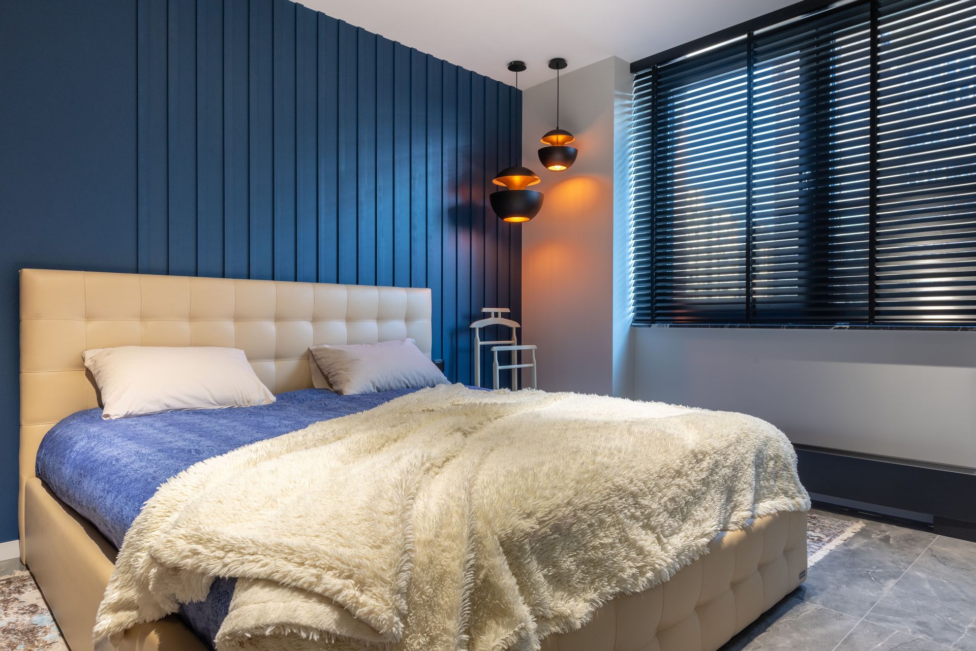 Modern bedroom with blue paneled wall, beige bed, cream throw, and dark blinds over a window