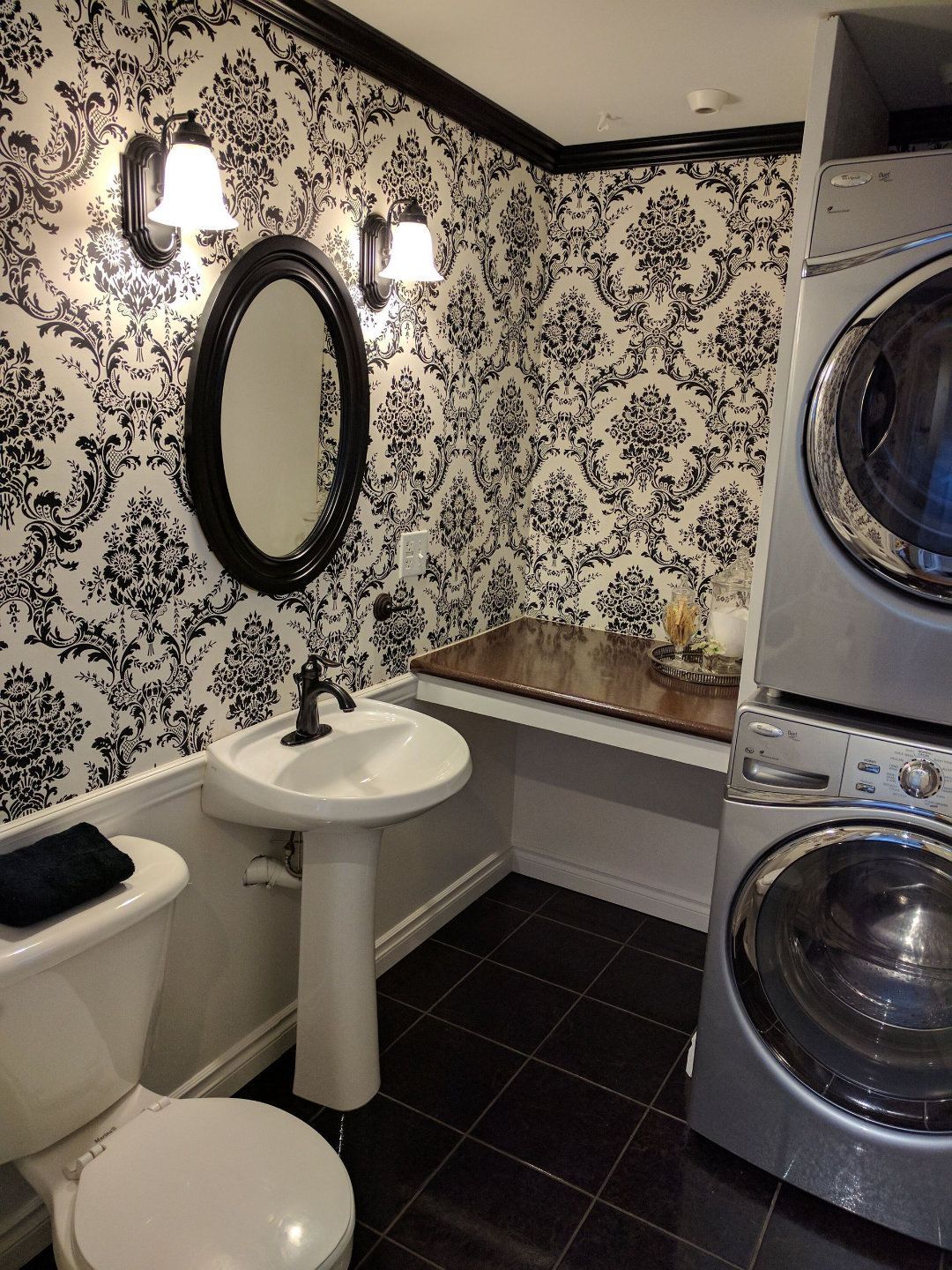 Compact laundry room with patterned wallpaper, pedestal sink, mirror, and stacked washer-dryer.