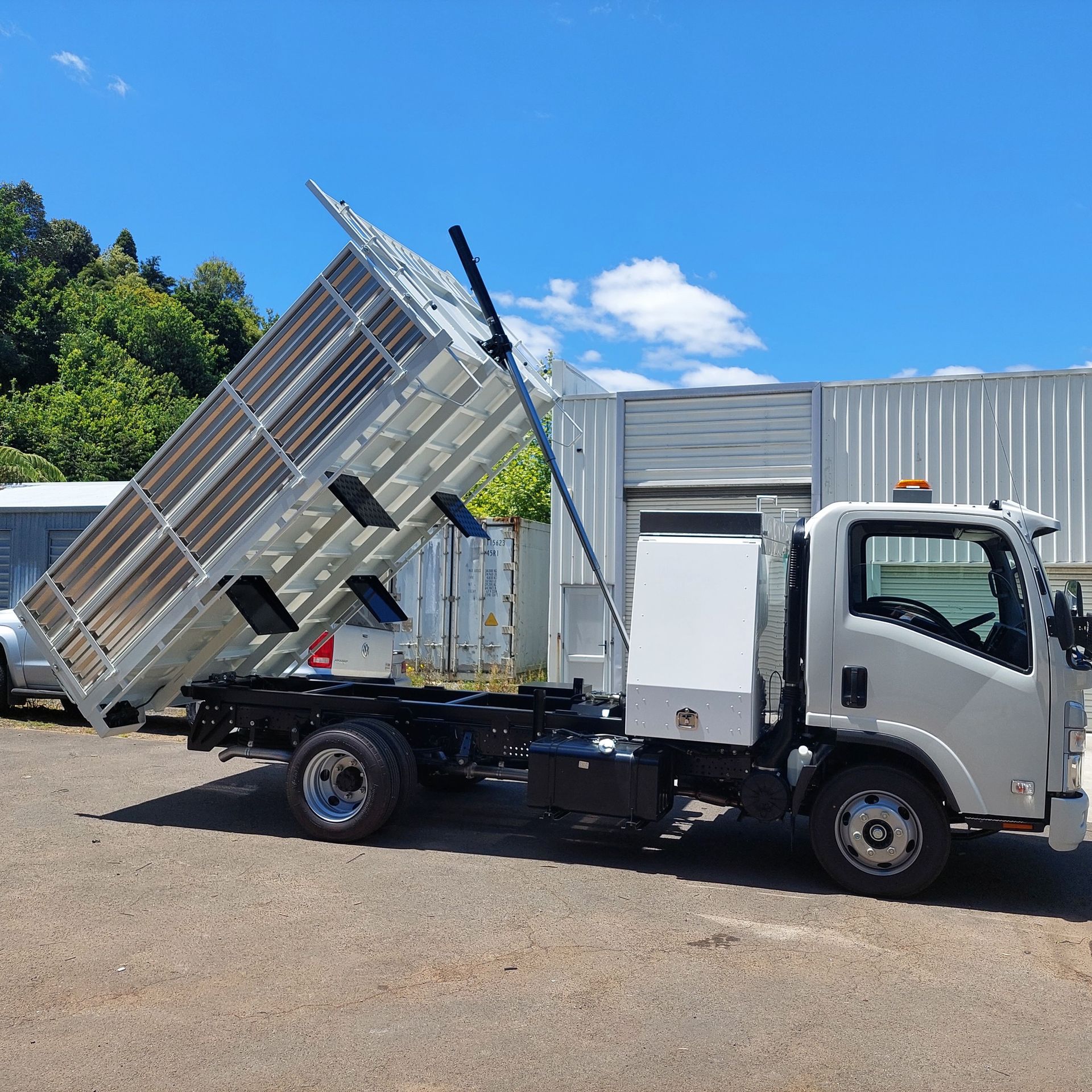 Tilted utility trailer with raised mesh ramp at an industrial site, attached to a truck.