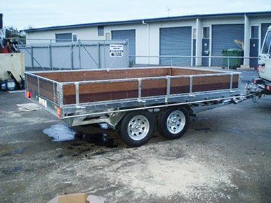 Flatbed utility trailer with metal rails parked in an industrial lot, with a white building in the background.