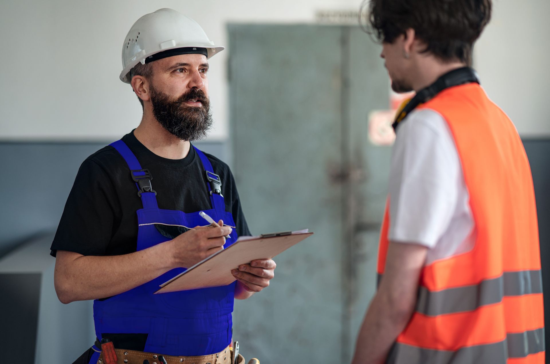 Two construction workers in hard hats, one writing on a clipboard while talking indoors.
