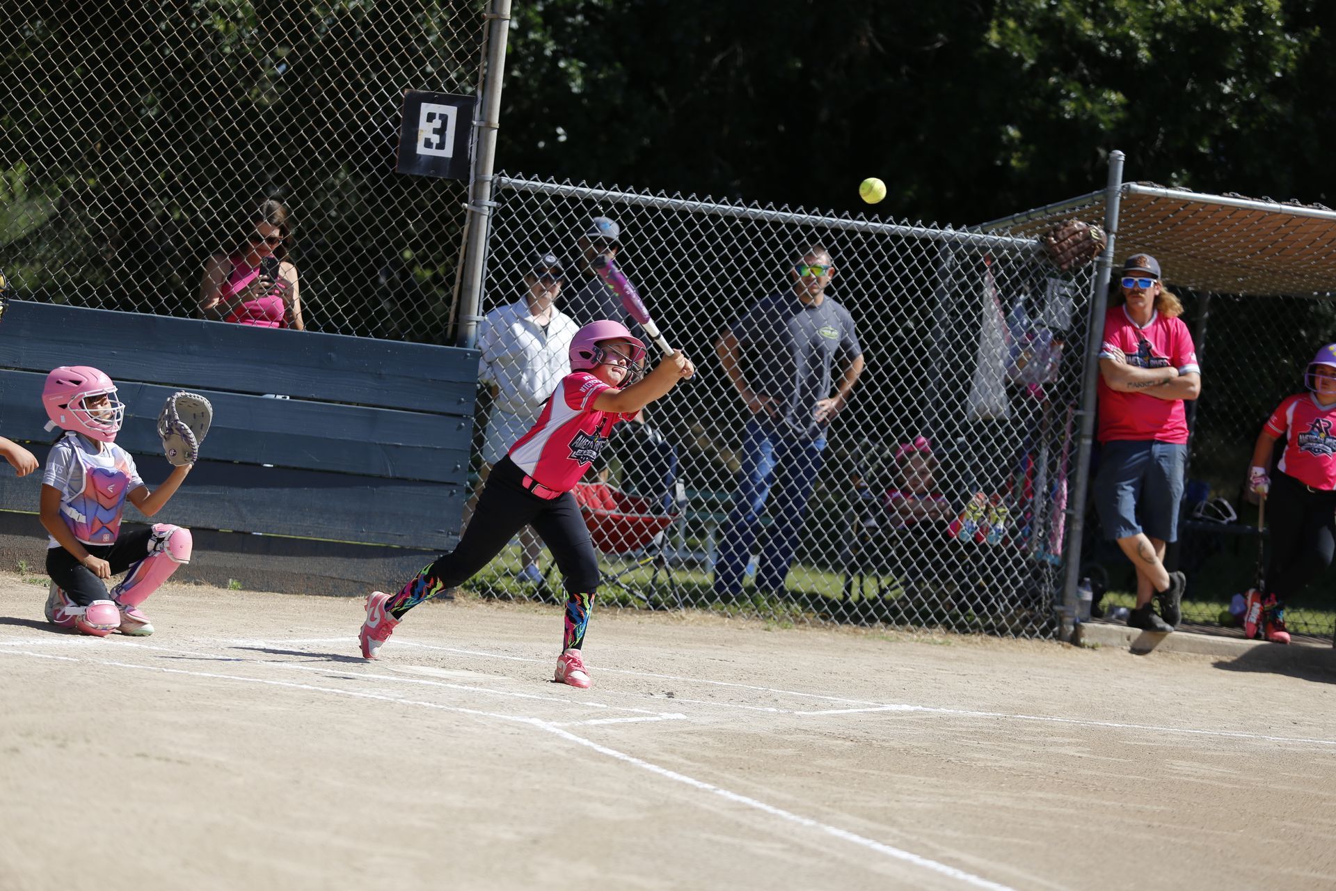 Softball player hitting the softball while the catcher, coaches and parents watch