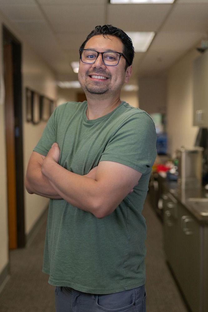 Smiling person in a green T-shirt standing with arms crossed in a hallway, with office lights behind them.