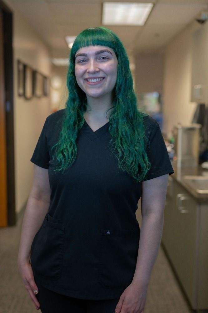 Person in black scrubs with long green hair smiling in a hallway.