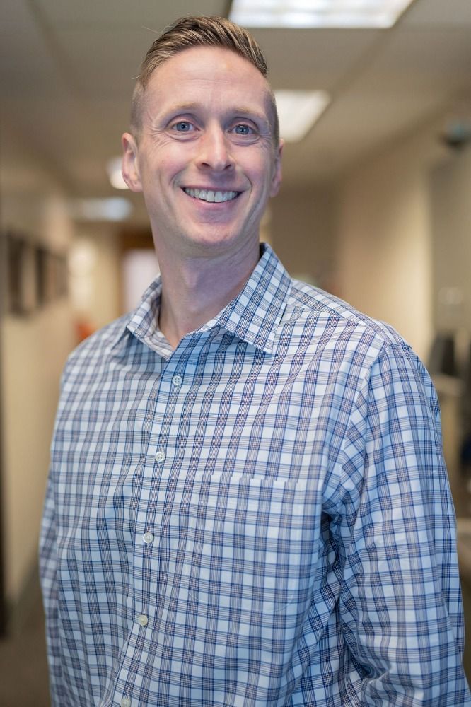 Smiling man in a blue plaid shirt standing in a hallway with a blurred background