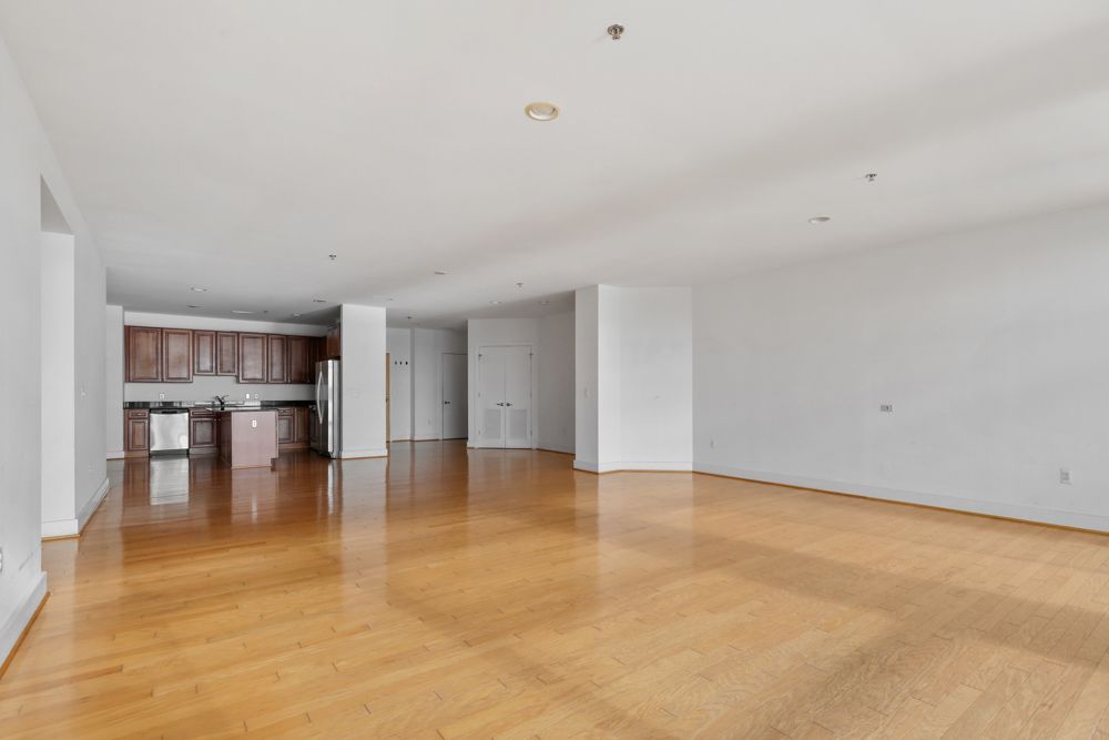 An empty living room with hardwood floors and a kitchen in the background.