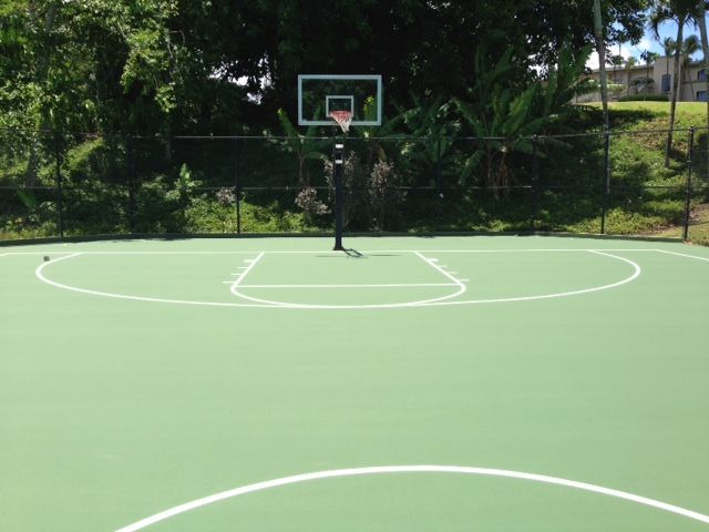Outdoor basketball court with blue borders, gray surface, and two hoops, set against a backdrop of trees and mountains.