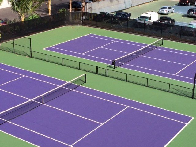 Two outdoor tennis courts, purple playing surface, green surround. Black nets and fencing. Cars parked behind.