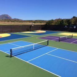Multi-sport court with blue, green, yellow, and purple surfaces. Nets for pickleball and basketball hoop are visible. Clear sky.