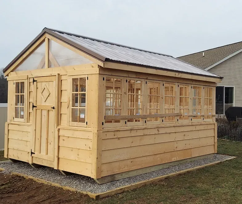 A wooden greenhouse with a metal roof is sitting in the dirt in front of a house.