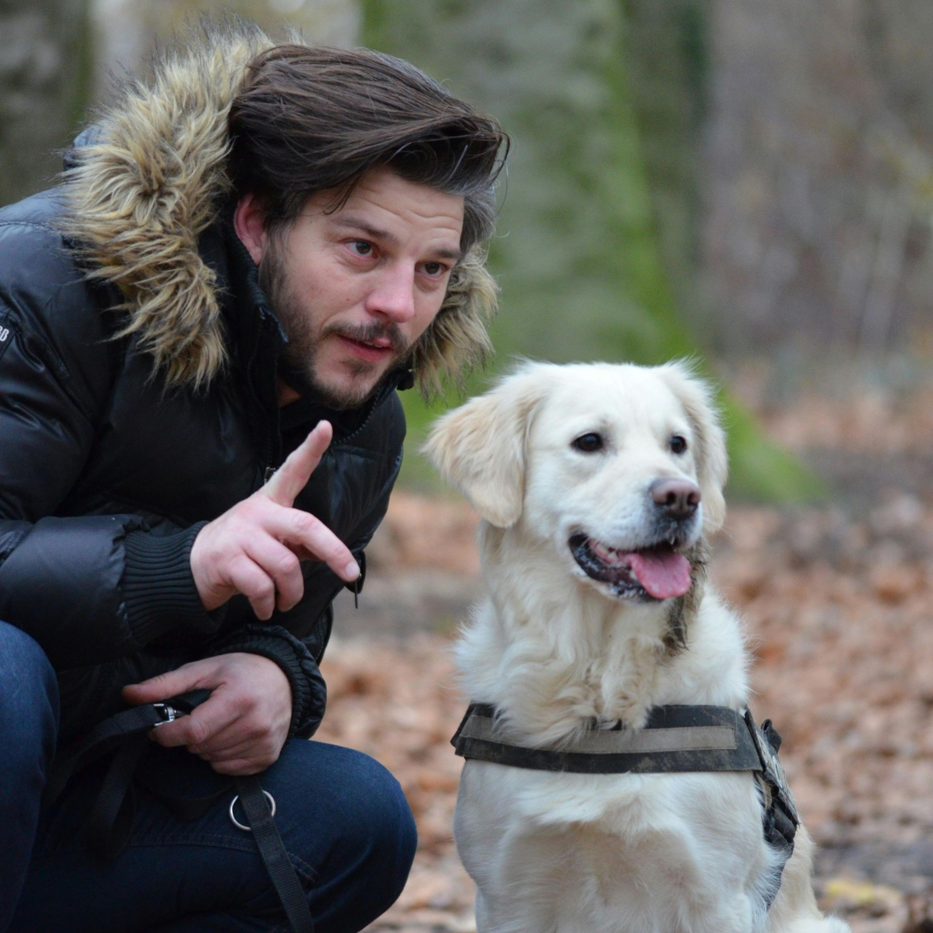 Man in winter coat pointing at golden retriever in forest.