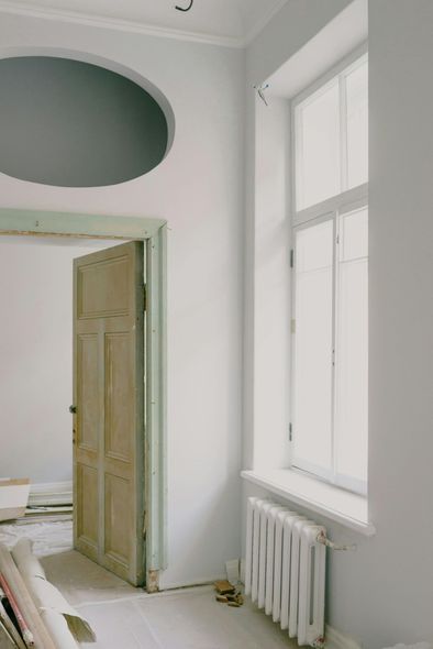 A room interior featuring a pale wood door, a tall window, a white radiator, and an oval cutout in the wall above the door.