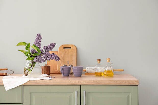 Kitchen counter with lilac bouquet, cutting boards, cups, oil bottles, and rolling pin. Green cabinets and gray wall.