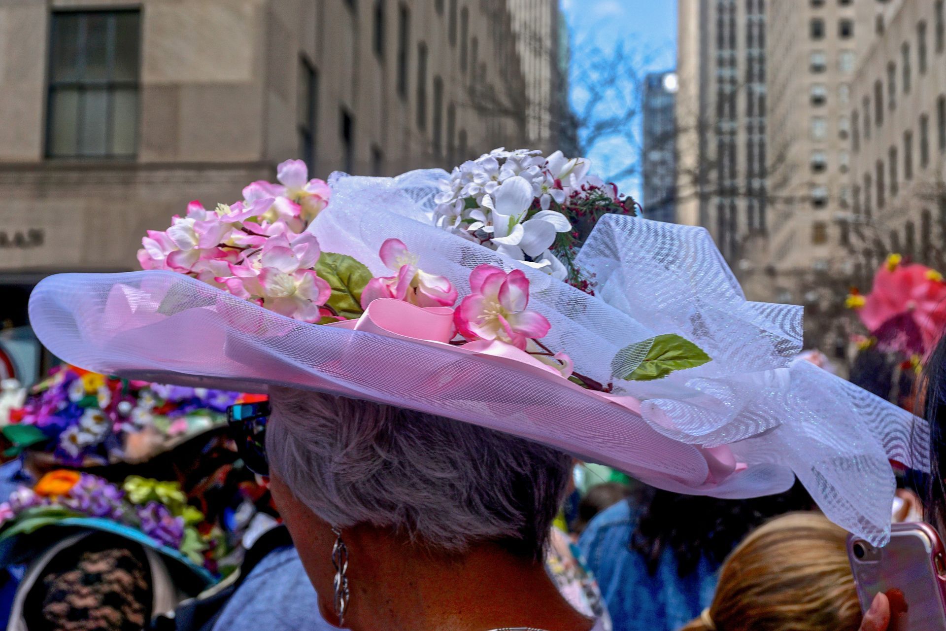 A person wears a large, wide-brimmed pink hat adorned with tulle and artificial flowers, viewed from behind in a city.