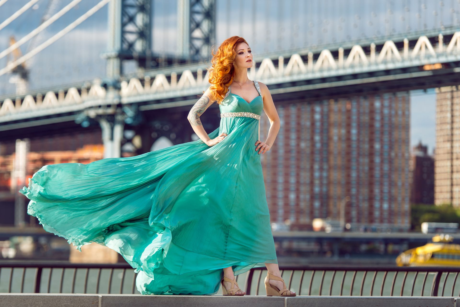 Woman in flowing turquoise dress poses near bridge and city skyline.