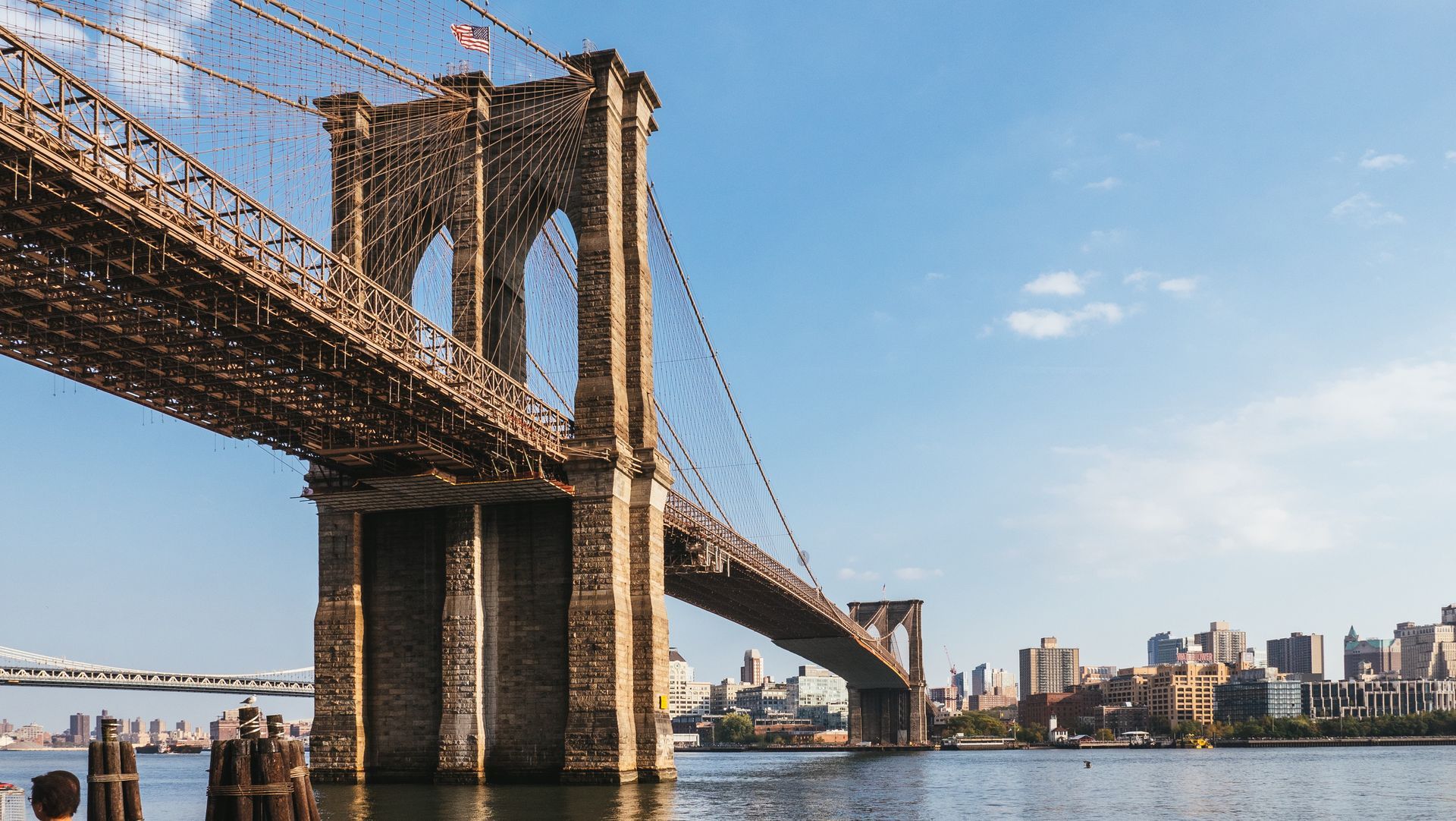 Brooklyn Bridge over water, cityscape in the background, blue sky.