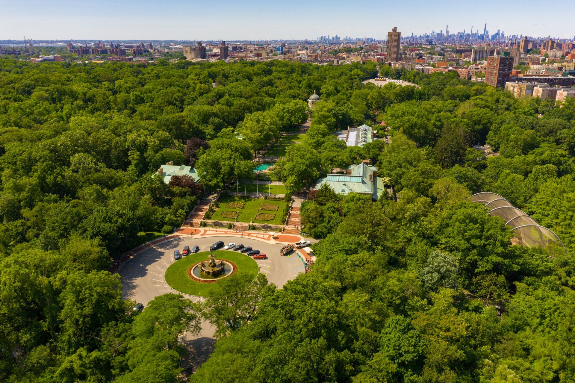 Aerial view of a lush green park with buildings, a circular driveway, and a city skyline in the distance.