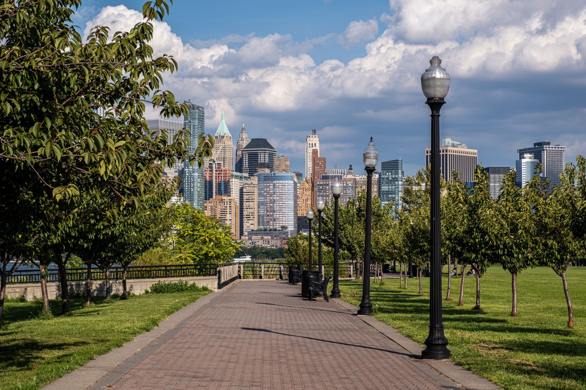 Brick path leading to a city skyline, lined with lampposts and trees, under a cloudy blue sky.