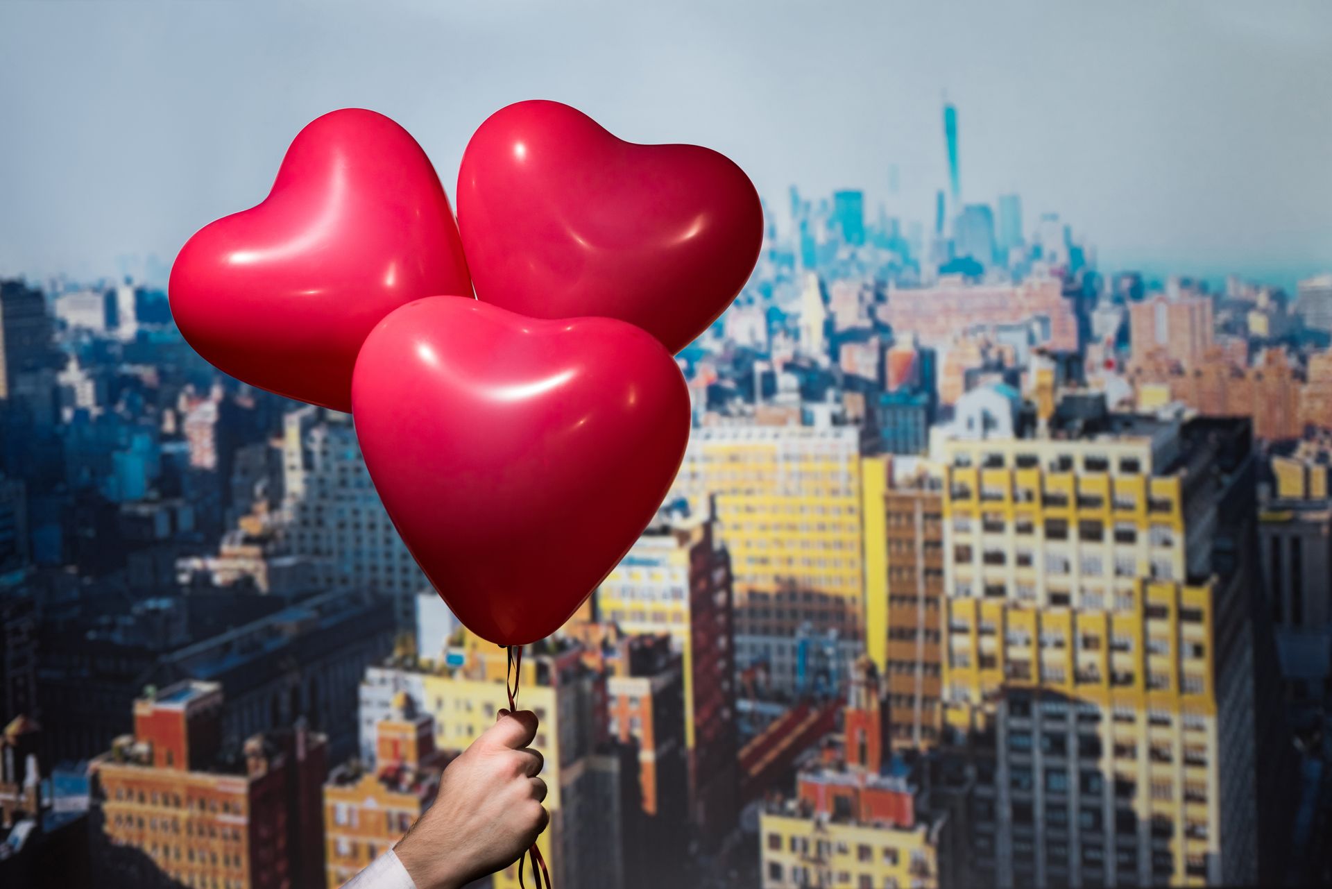 Hand holding three red heart-shaped balloons against a New York City skyline.