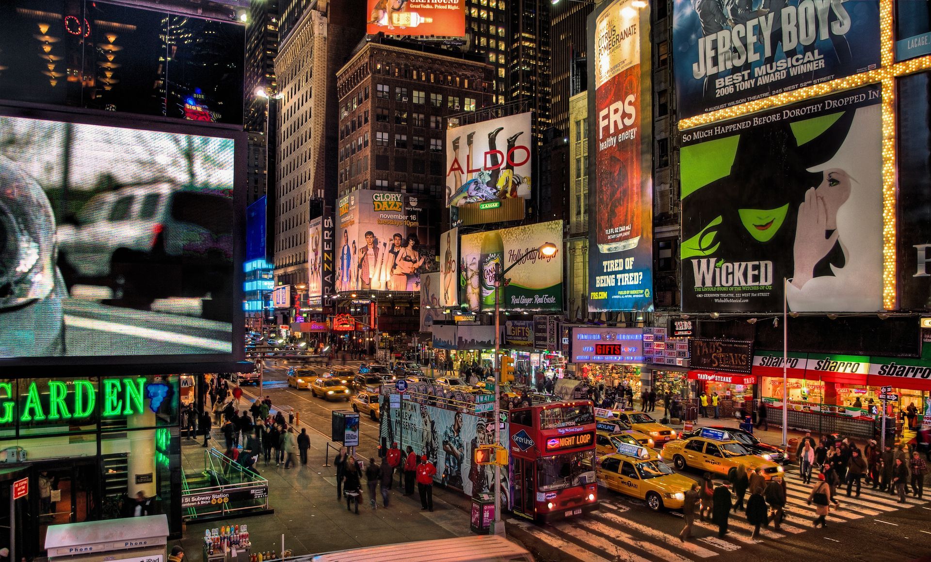 Times Square at night, brightly lit with billboards, crowds, taxis, and a double-decker bus.