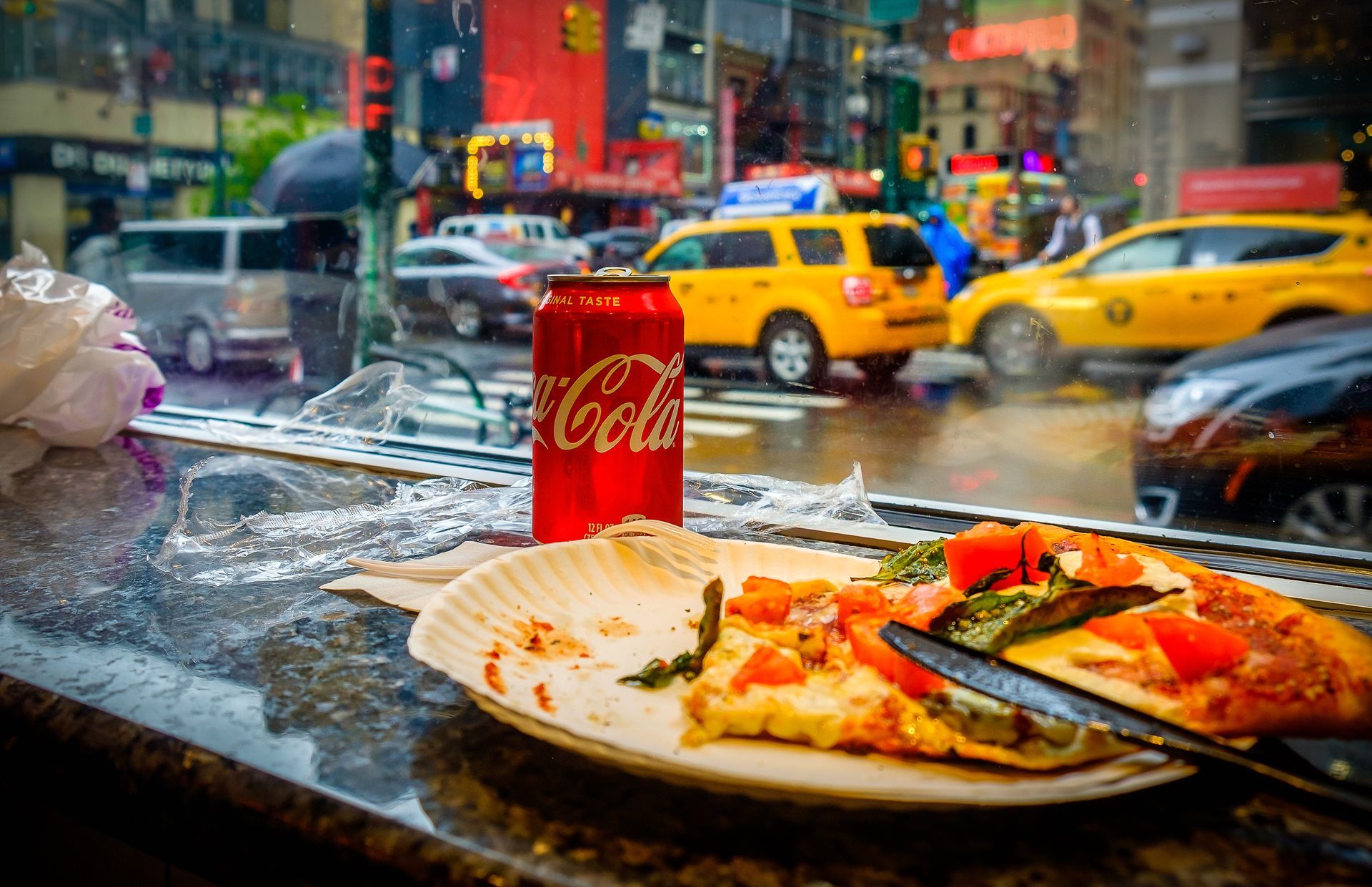 A slice of pizza and a can of coca cola are on a table in front of a window.
