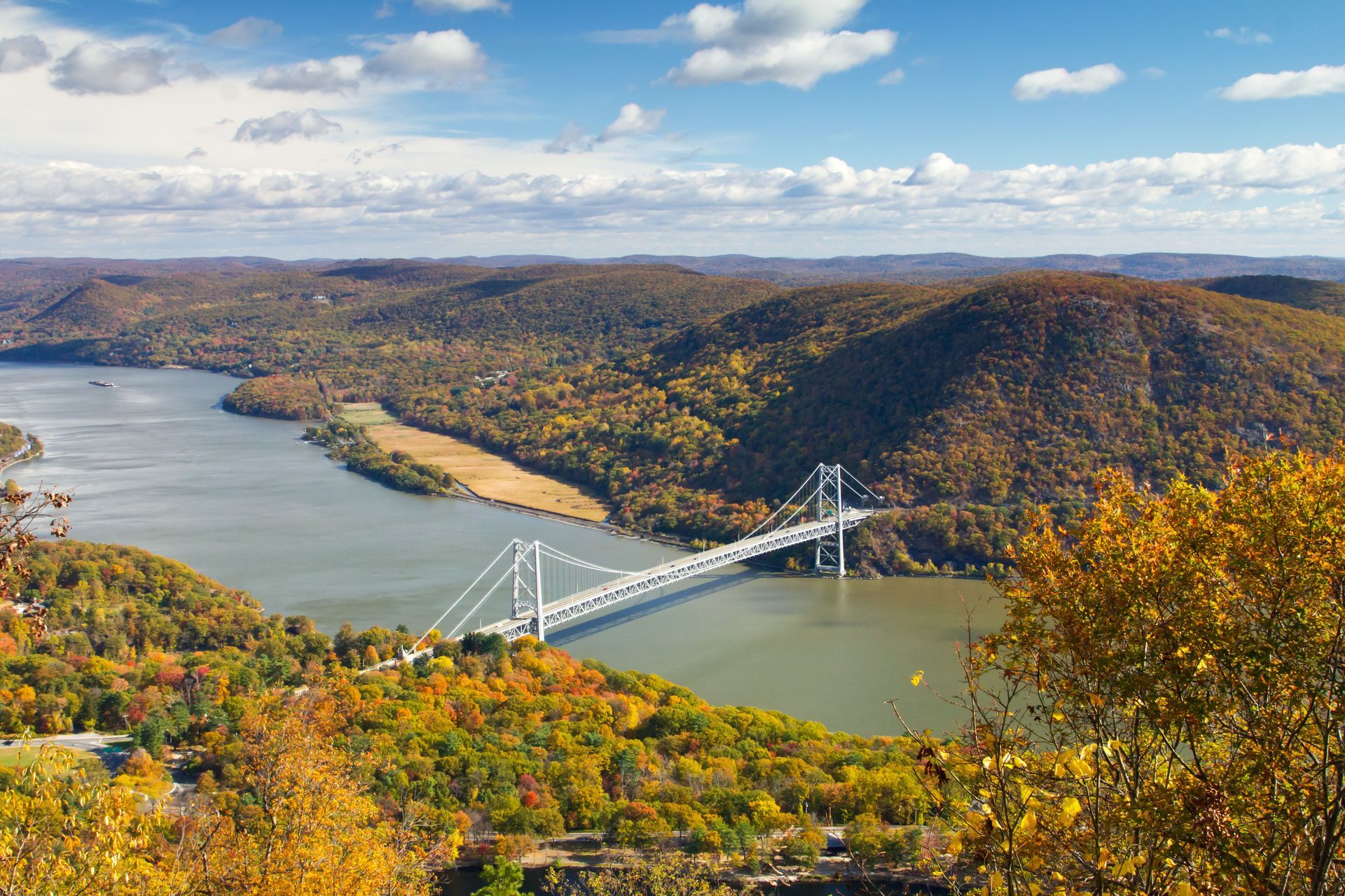 Suspension bridge spanning a wide river, surrounded by hills covered in autumn foliage. Blue sky with clouds.