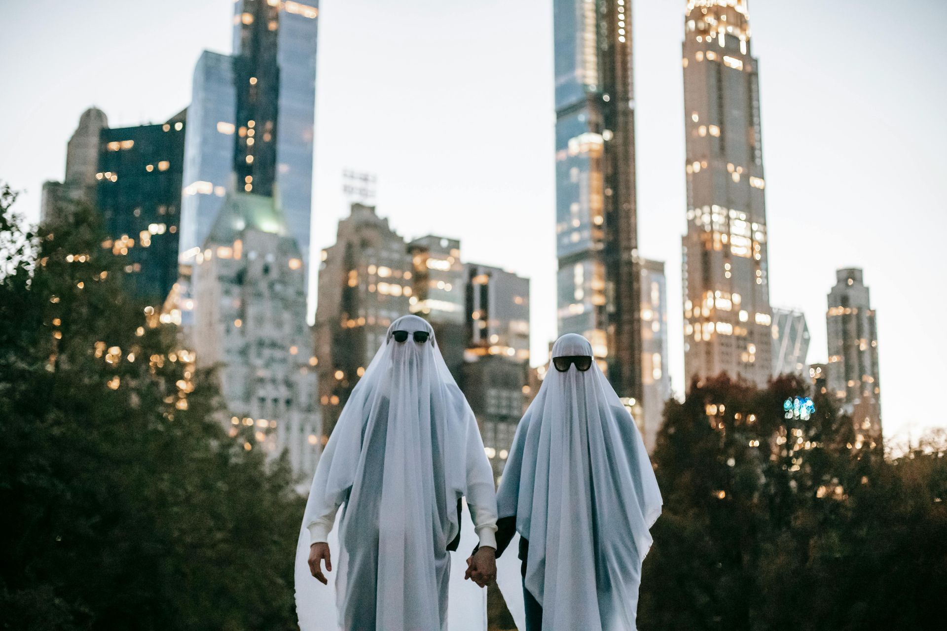 Two people in ghost costumes, holding hands, with sunglasses on, in front of a cityscape.