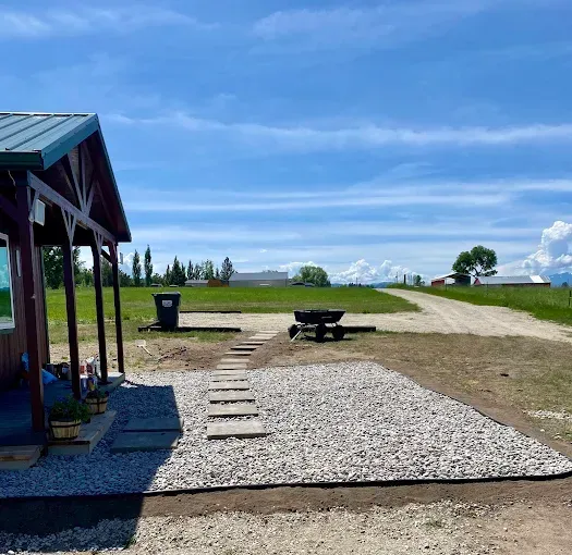 A gravel patio and stone walkway lead away from the porch of a cabin into a large, sunny field under a blue sky.