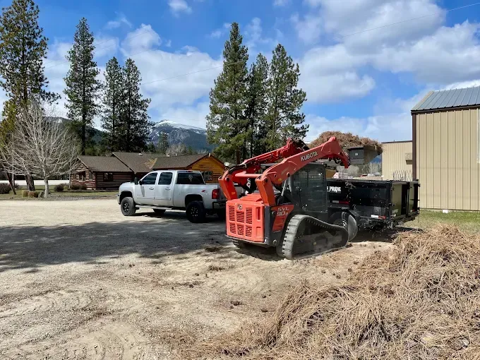 An orange skid steer loader parked next to a black dump trailer in a gravel lot with a white truck and mountains behind.