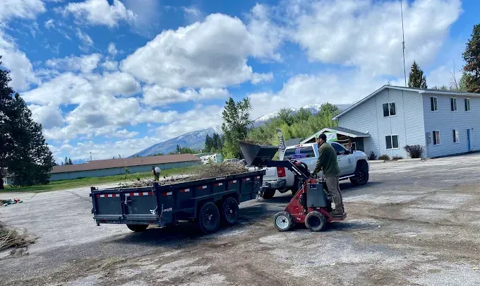 A person operates a red skid steer to load debris into a black dump trailer parked next to a white truck near a building.