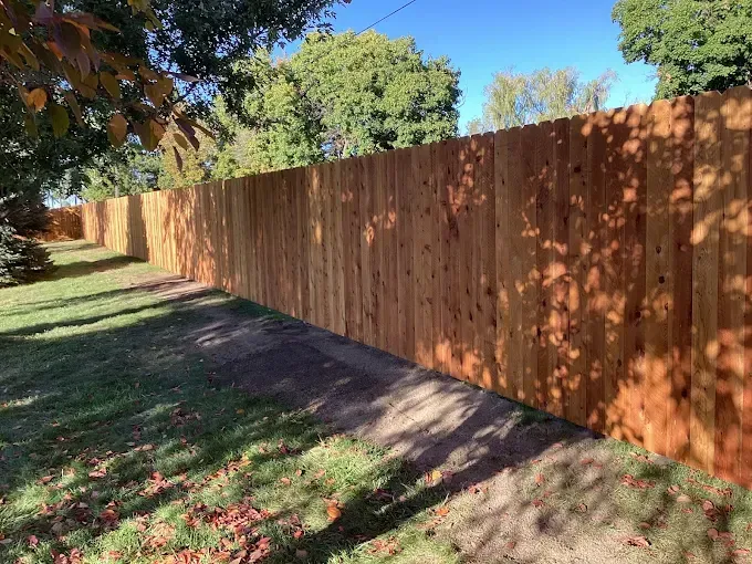 A long, wooden privacy fence stands along a grassy lawn on a sunny day with trees in the background.