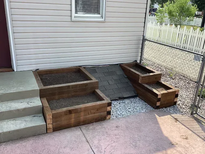 Two tiered wooden garden beds flanking a basement window well against a house with light siding.