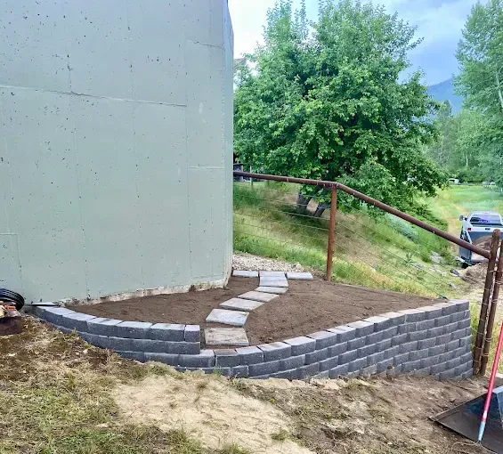 A curved grey retaining wall built of stone blocks wraps around a dirt patch with a small stone path by a large tank.