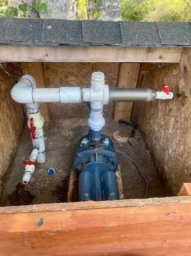 A water pump with connected PVC piping and red-handled shut-off valves inside a small, wooden enclosure.