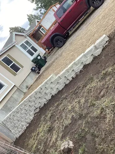 A red pickup truck is parked on a gravel driveway next to a light-colored house with a stacked stone retaining wall.