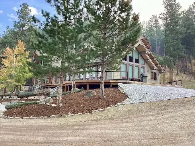 A modern A-frame house with a wooden deck nestled in a pine forest, featuring a gravel driveway and landscaped mulch.