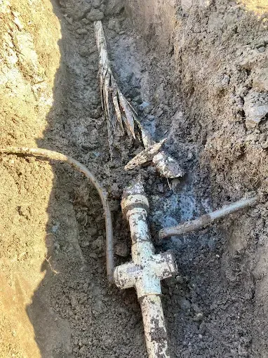 A damaged, white utility pipe with a four-way connector lies exposed in a dirt trench, surrounded by mud and debris.