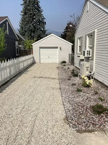 A gravel driveway leads to a detached white garage between a house with a white fence and a home with light siding.