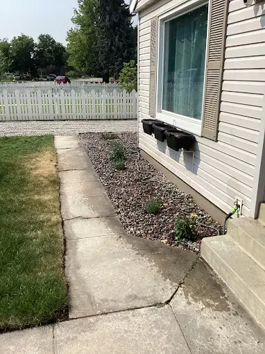 A concrete walkway leads past a house exterior with black window flower boxes and a rock garden bed alongside a lawn.