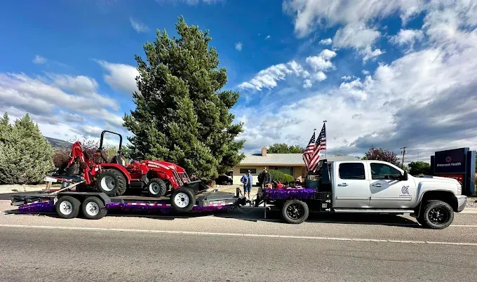 A white pickup truck with American flags towing a trailer carrying a red tractor on a sunny day.
