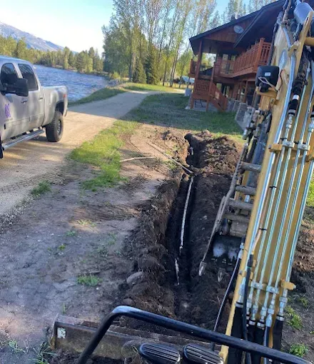 Excavator digging a trench alongside a rural road and building near a lake.