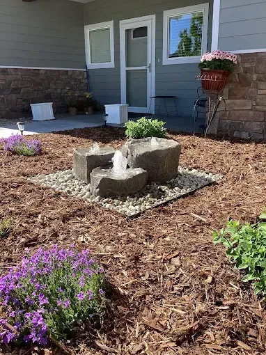 A stone fountain flows in a front yard garden with brown mulch, river rocks, and purple flowers near a grey house.