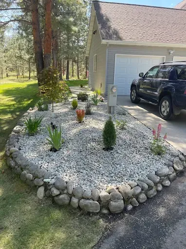 A rock-bordered garden bed with small shrubs and plants in front of a house garage with a dark SUV parked on the driveway.