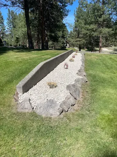 A long, narrow rock garden with a concrete border, situated in a grassy lawn area surrounded by pine trees.