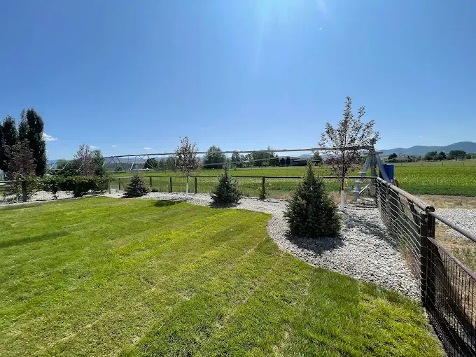 A mowed green lawn borders a gravel bed with small pine trees and a fence, overlooking a sunlit field under a blue sky.