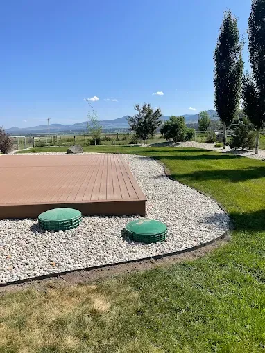 A brown deck with two green septic access covers in a gravel border, set in a sunny backyard with trees and mountains.