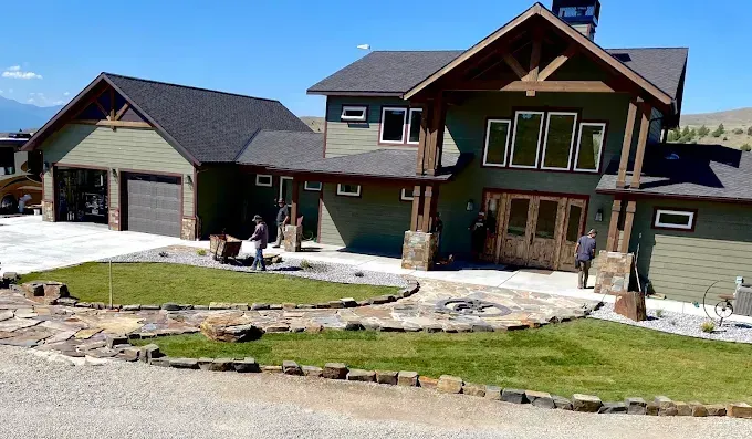 Workers landscaping the front yard of a two-story green rustic home with stone accents and a gravel driveway.